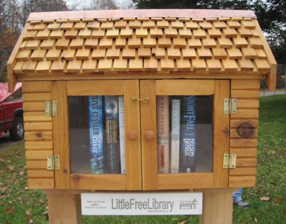 A wooden little free library with a shingled roof and glass doors, containing several books.